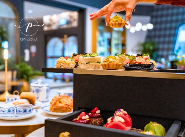 A hand reaching for a delicious mini tart at the afternoon tea service at Prologue in The Dorian Hotel, Calgary, showcasing an array of vibrant, gourmet finger foods.